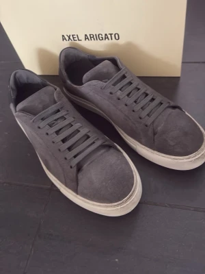 Gråa Axel Arigato sneakers i mocka - Snygga gråa sneakers från Axel Arigato i mjuk mocka med diskret guldfärgad logga på sidan. Klassisk låg modell med vita sulor och matchande grå skosnören. Perfekta för en clean och stilren look.