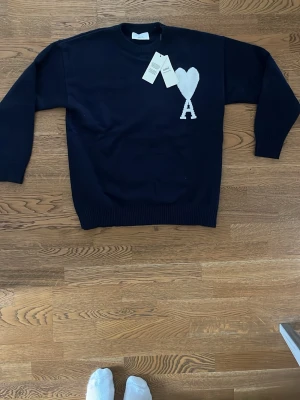 Svart stickad tröja från Ami Paris - Svart stickad tröja från Ami Paris med vit hjärtlogga och bokstaven A på bröstet. Klassisk rund halsringning och ribbstickade muddar vid ärmslut och nederkant. Perfekt för en stilren och trendig look.