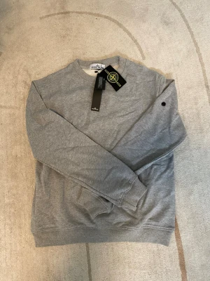 Grå sweatshirt från Stone Island - Säljer en stilren grå sweatshirt från Stone Island med klassisk rund hals och ribbade muddar. Tröjan har lång ärm och den ikoniska Stone Island-loggan på ärmen. Perfekt till ett avslappnat streetwear-look.