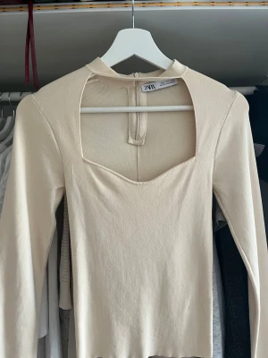 Beige ribbad topp med cut out från Zara - Trendig beige långärmad topp från Zara med ribbad struktur och unik cut out-detalj vid halsen. Toppen har en smickrande passform och är perfekt för dig som vill sticka ut lite extra. Supersnygg att styla med både jeans och kjol.