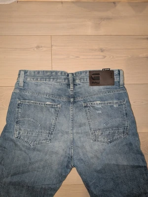 Ljusblå jeans från G-Star RAW - Säljer ett par ljusblå jeans från G-Star RAW med klassisk femficksmodell och raka ben. Jeansen har slitna detaljer och en snygg läderpatch med logga bak i midjan. Tillverkade i denim med en avslappnad passform, perfekta för en casual look. Helt nya