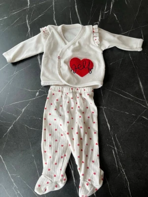 Vitt babyset med röda hjärtan - Sött tvådelat babyset i vitt med röda hjärtan. Tröjan har långa ärmar, volangdetaljer på axlarna och ett rött hjärta med texten 'self' broderat framtill. Byxorna har resår i midjan och små röda hjärtan över hela tyget. Perfekt för mysiga dagar.