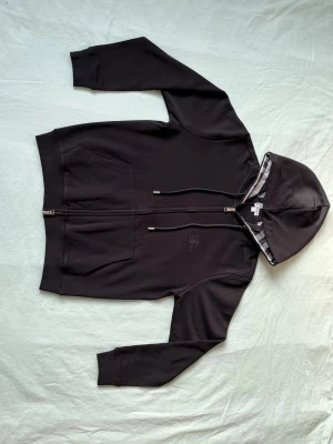 Svart hoodie från Burberry med dragkedja - Svart hoodie från Burberry med dragkedja framtill och klassisk logga broderad på bröstet. Huvan har ett snyggt rutigt foder och justerbara snören med metalländar. Tillverkad i mjuk bomull för en skön känsla. Perfekt för en stilren och avslappnad look.