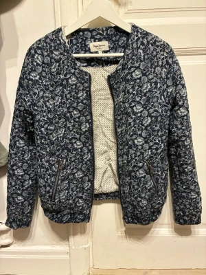 Blommig cardiganjacka från Pepe Jeans - Mönstrad cardiganjacka från Pepe Jeans i marinblått med vita blommor. Jackan har rund halsringning, dragkedjefickor och elastisk nederkant. Insidan är ljus med diskret prickmönster. Perfekt för dig som gillar en unik och trendig stil.