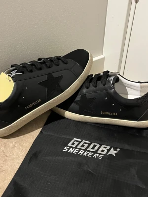 Svarta GGDB Superstar sneakers - Snygga svarta GGDB Superstar sneakers med svart stjärndetalj på sidan, svarta skosnören och vit/gräddfärgad sula. Ovandelen är i läder och har GGDB/SSTAR-tryck på sidan. Klassisk låg modell med retrovibbar och slitstark yttersula.