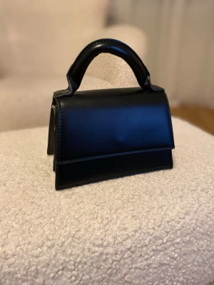Svart minihandväska i läderlook från NK-AD - Stilren svart minihandväska med kort handtag och elegant, kantig form. Väskan har en slät yta i läderimitation och minimalistisk design utan synliga loggor. Perfekt accessoar för att lyfta din outfit. Från NK-AD den är bara använd en gång 
