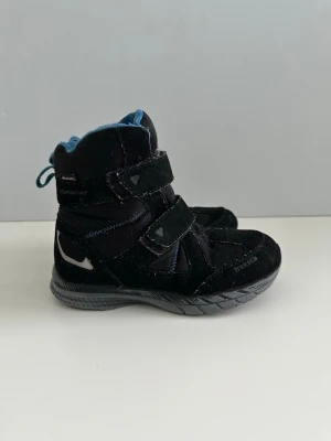 Svarta sneakers från Treksta, storlek 32 - Säljer ett par svarta sneakers från Treksta med blått foder och två kardborreband för enkel påtagning. Skorna har robust sula och detaljer i reflex samt mockaliknande material. Perfekta för dig som vill ha både stil och funktion.