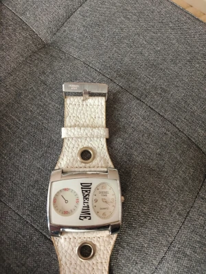 Diesel vit klocka med bred rem - Snygg klocka från Diesel med bred, vit rem och silverfärgat spänne. Urtavlan är rektangulär med två separata visningsfönster och Diesel-logga i mitten. Klockan har en unik design och detaljerad struktur på remmen.