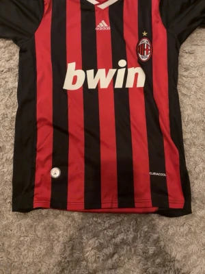AC Milan Ronaldinho matchtröja - Säljer en klassisk AC Milan matchtröja från Adidas med Ronaldinho och nummer 80 på ryggen. Tröjan har röda och svarta vertikala ränder, klubbmärke på bröstet och Bwin som sponsor. Tillverkad i lätt och ventilerande ClimaCool-material.