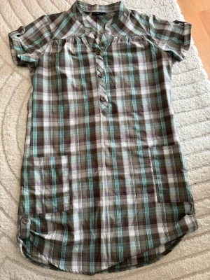 Rutig kort klänning från Lindex - Kort skjortklänning från Lindex i rutigt mönster med bruna, turkosa och vita toner. Klänningen har korta ärmar, knappar framtill och en avslappnad passform. Perfekt för varma dagar och enkel att styla med sneakers eller sandaler.