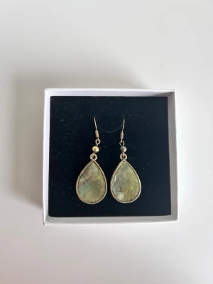 Droppformade örhängen med grön sten - Snygga droppformade örhängen med en grön skimrande sten och guldfärgade detaljer. Örhängena har en elegant och enkel design med krokar för enkel användning. Perfekta för att ge din look en unik touch.