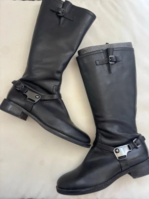 Svarta läderstövlar med spännen - Snygga svarta stövlar i äkta läder med dekorativa spännen både upptill och vid ankeln. Stövlarna har en robust klack och metall-detaljer som ger en cool look. Perfekta för dig som vill klä upp en outfit.