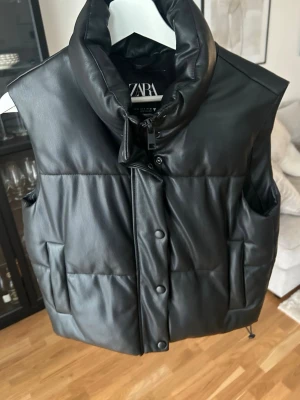Svart puffer-väst i fejkläder från Zara - Säljer en svart puffer-väst från Zara i fejkläder med hög krage och tryckknappar framtill. Västen har två sidofickor och en oversized passform. Perfekt för lager på lager.💓
