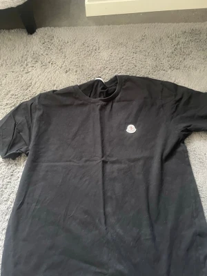 Svart Moncler t-shirt i bomull - Snygg svart t-shirt från Moncler med klassisk rund hals och korta ärmar. På bröstet sitter Monclers ikoniska logotyp i vitt och rött. Tillverkad i mjuk bomull som känns skön mot huden. Perfekt för en clean och stilren look.