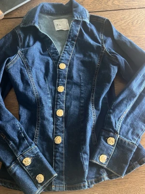 Mörkblå jeansjacka XS med guldknappar - Snygg mörkblå jeansjacka från Zara i storlek XS. Figurnära modell med markerade sömmar och stora guldiga knappar framtill och vid ärmsluten. Klassisk krage och långa ärmar. Perfekt för dig som gillar en stilren och trendig look.