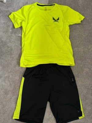 Neongult träningsset från SOC - Säljer ett sportigt träningsset från SOC med en neongul t-shirt i funktionsmaterial och matchande svarta shorts med neongula detaljer på sidorna. Perfekt för gymmet eller löprundan. Materialet är lätt och andas bra.