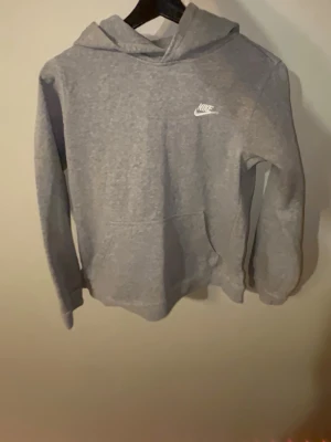 Grå Nike hoodie med logga - Snygg grå hoodie från Nike med klassisk vit logga på bröstet. Hoodien har huva och en stor magficka framtill. Tillverkad i mjukt material som är skönt att ha på sig. Perfekt för en avslappnad och sportig stil.