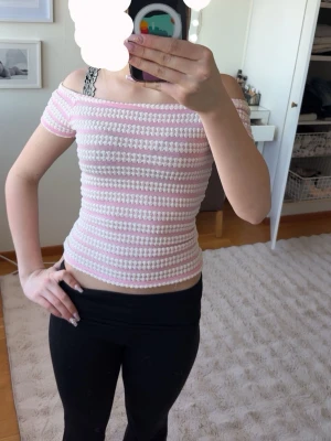 Randig offshoulder topp i rosa och vitt - Söt offshoulder topp med korta ärmar i stickat material. Toppens mönster består av ljusrosa och vita ränder, och den är croppad med en slim passform. Perfekt för dig som gillar en feminin och trendig stil.