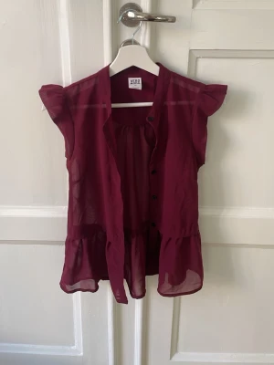 Vinröd blus från Vero Moda - Snygg vinröd blus från Vero Moda i transparent, lätt material. Blusen har volangdetaljer nedtill och vid axlarna samt knappar framtill.