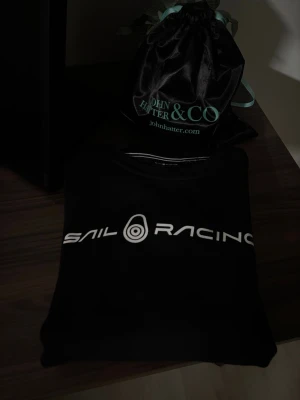 Svart tröja från Sail Racing - Snygg svart tröja från Sail Racing med vit logotyp tryckt över bröstet. Tröjan har rund halsringning och är tillverkad i mjukt material, perfekt för en avslappnad och väldigt grisch