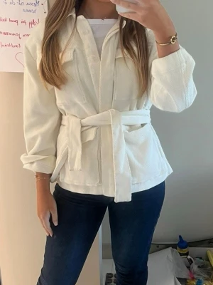Vit overshirt med knytskärp - Stilren vit overshirt i mjukt tyg med knytskärp i midjan och två bröstfickor. Klassisk krage och knäppning framtill. Perfekt att slänga över en t-shirt för en avslappnad men trendig look.