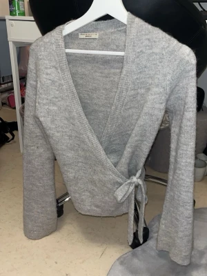 Grå omlottkofta från Gina tricot - Mysig grå omlottkofta från Gina tricot  med knytband i midjan och djup V-ringning. Koftan har långa ärmar och är stickad i ett mjukt material som passar perfekt för lager på lager. Enkel och stilren design som funkar till det mesta.
