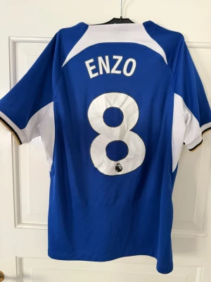 Chelsea matchtröja Enzo #8 Nike - Säljer en blå Chelsea matchtröja med Enzo och nummer 8 på ryggen. Tröjan har Premier League-märke på ärmen, sponsorlogga 'INFINITE ATHLETE' på bröstet och klubbmärke fram. Tillverkad i lätt och ventilerande material, perfekt för fotboll eller träning.(hemmatröja 23/24