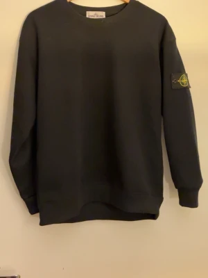 Svart sweatshirt från Stone Island - Klassisk svart sweatshirt från Stone Island med rund halsringning och ribbade muddar. Tröjan har den ikoniska Stone Island-loggan på vänster ärm. Perfekt för en avslappnad och stilren look.