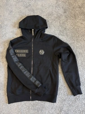 Svart hoodie från Philipp Plein - Svart hoodie från Philipp Plein med dragkedja framtill och huva. Märkets logga och namn syns tydligt på bröstet och längs ena ärmen. Hoodien har fickor framtill och är tillverkad i mjuk bomull för en bekväm känsla.