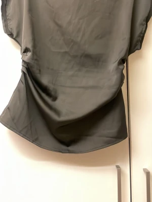 Svart satängblus med korta ärmar - Stilren svart blus i satin  satängliknande material med lös passform och korta ärmar. Perfekt att matcha med jeans eller kjol för en trendig look. Blusen har rund halsringning och lätt drapering i sidorna.