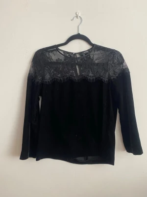 Svart topp med spetsdetaljer - Snygg svart topp med lång ärm och transparent spets upptill över axlar och bröst. Toppen har rund halsringning och knapp i nacken. Materialet är mjukt och känns lyxigt, perfekt för att lyfta vilken outfit som helst.