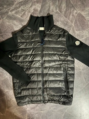 Moncler  - Snygg svart dunväst från Moncler med stickade svarta ärmar och hög krage. Västen har två dragkedjeförsedda fickor framtill och ett Moncler-märke på ena ärmen. Perfekt för lager-på-lager och riktigt trendig look.
