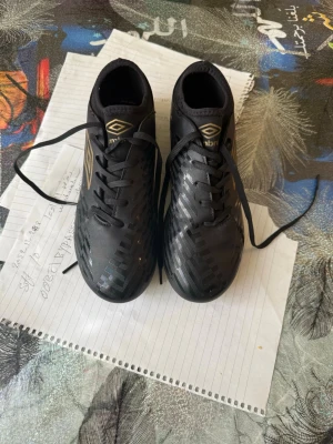 Svarta Umbro fotbollsskor - Säljer ett par svarta fotbollsskor från Umbro med guldfärgade loggor på sidan och tungan. Skorna har snörning och mönstrad ovandel i syntetmaterial för bra bollkontroll. Perfekta för dig som vill ha stilrena och funktionella skor på planen.