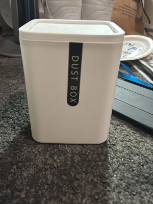Vit liten papperskorg Dust Box - Säljer en vit, rektangulär papperskorg med texten 'DUST BOX' på framsidan. Tillverkad i plast och har lock. Perfekt för skrivbordet eller små utrymmen.