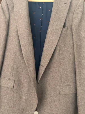 Brun kavaj med mönstrat foder - Stilren brun kavaj med klassisk passform och två fickor framtill. Insidan har ett mörkblått foder med små orange och röda sköldmönster. Perfekt för dig som vill ha en tidlös look med en twist. Materialet är ullblandning. Säljes till det löjligt låga priset 550kr