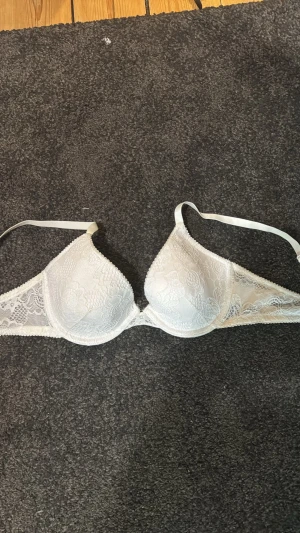 Vit spets-bh med bygel - Säljer en vit bh med bygel och vacker spetsdetalj. Bh:n har justerbara axelband och klassisk knäppning bak. Materialet är mjukt och spetsen ger en elegant look. Perfekt för dig som gillar stilrena underkläder med lite extra detaljer.