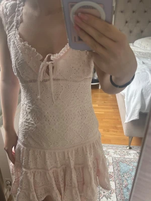 Ljusrosa spetsklänning med volang - Söt ljusrosa kortklänning med spetsdetaljer och volangkant. Klänningen har breda axelband, omlott-ringning med snörning framtill och ett romantiskt mönster. Perfekt för sommarens alla tillfällen. Endast använd en gång o säljer då den är för stor 