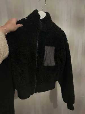 Svart teddyjacka med dragkedja - Mysig svart teddyjacka med en hel dragkedja framtill. Jackan har en grå bröstficka med dragkedja och ribbade muddar vid ärmslut och nederkant