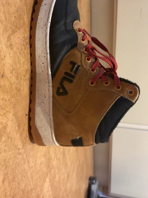 Bruna och svarta kängor från Fila - Snygga kängor från Fila i brunt och svart med röda snören och detaljer i gummi. Ovandelen är i mocka och textil, med robust sula och högt skaft. Perfekta för dig som vill ha en streetstyle-vibe och gillar sportiga detaljer.