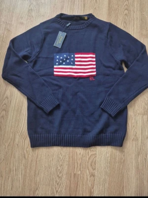 Mörkblå stickad tröja Ralph Lauren - Mörkblå stickad tröja från Ralph Lauren med amerikansk flagga broderad på bröstet och RL-bokstäver. Tröjan har rund hals, långa ärmar och ribbstickade muddar. Perfekt för dig som gillar klassisk och stilren design.