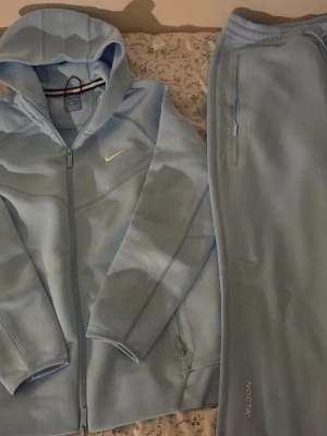 Ljusblå Nike NOCTA tracksuit - Säljer ett ljusblått tracksuit-set från Nike NOCTA med hoodie och matchande byxor. Hoodien har dragkedja, fickor och NOCTA-detaljer, samt Nike Swoosh på bröstet. Byxorna har dragkedjeficka och NOCTA-logga. Perfekt för sport eller chill. Materialet är mjukt och stretchigt.