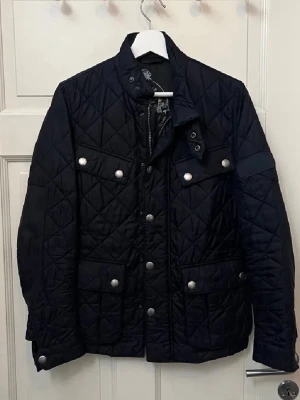 Svart quiltad jacka från Barbour - Snygg svart quiltad jacka från Barbour med silverfärgade knappar och dragkedja. Jackan har flera fickor framtill, hög krage och ett coolt innerfoder med tryck. Perfekt för dig som gillar klassisk men modern stil.