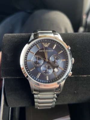 Emporio Armani silver herrklocka - Stilren herrklocka från Emporio Armani med silverfärgad länkarmband och mörkblå urtavla. Klockan har kronograf-funktion, datumvisning och tydliga indexmarkeringar. Perfekt accessoar för dig som gillar klassisk och modern stil.