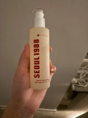 K-Secret Seoul 1988 Essence Toner - K-Secret Seoul 1988 Essence Toner med 97% snigelsekret och risextrakt. Kommer i en genomskinlig plastflaska med pump och röd text. Perfekt för dig som vill ha återfuktad och fräsch hud med koreansk hudvård.