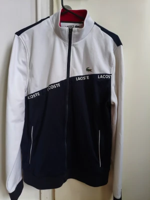 Vit och marinblå ziptröja Lacoste - Snygg ziptröja från Lacoste i vitt och marinblått med hög krage och dragkedja framtill. Tröjan har Lacoste-logga på bröstet och texten 'Lacoste' längs bröstet. Två fickor framtill och sportig design.