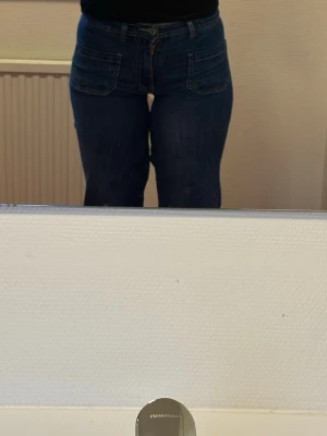 Mörkblå bootcut jeans med fickor - Säljer ett par mörkblå bootcut jeans med stora framfickor och markerade sömmar. Jeansen har låg midja och vida ben, vilket ger en avslappnad men trendig look. Perfekta att matcha med crop tops eller skjortor för en cool stil.