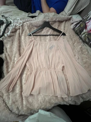 Ljusrosa plisserad blus med volang - Söt ljusrosa blus med plisserat tyg och volang nedtill. Blusen har tunna axelband och vida, långa ärmar som ger en luftig och romantisk känsla. Perfekt att styla med jeans eller kjol för en trendig look.