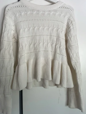 Vit stickad volangtröja Gina Tricot - Supersöt vit stickad tröja från Gina Tricot med volang nedtill och kabelstickat mönster. Tröjan har rund halsringning, långa ärmar och ribbade muddar. Perfekt för dig som gillar en feminin och trendig look.