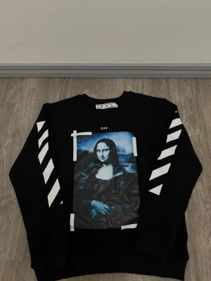 Off-White svart sweatshirt Mona Lisa - Svart sweatshirt från Off-White med ikoniskt Mona Lisa-tryck i blå toner på bröstet och vita diagonala ränder längs båda ärmarna. Rund halsringning och ribbade muddar. Perfekt för dig som gillar streetwear och vill sticka ut.