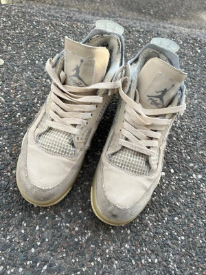  Off-White x Air Jordan 4  - Säljer ett par beige Off-White x Air Jordan 4 sneakers med meshdetaljer, genomskinliga plastpaneler och texten 'AIR' på sulan. Skorna har högt skaft, klassiska snörningar och ikoniska Jumpman-loggan på plösen. Perfekta för dig som gillar streetwear och unika samarbeten.